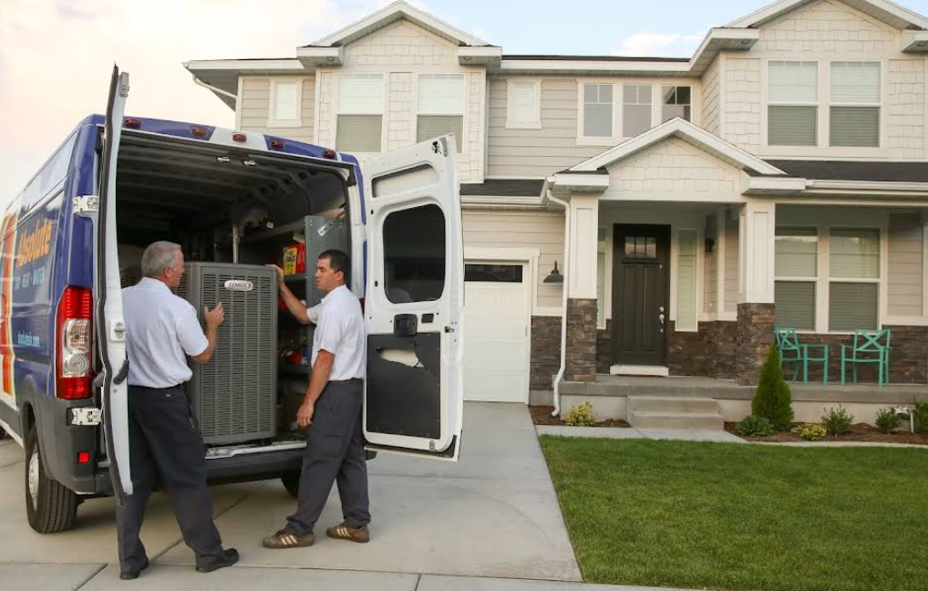 Two HVAC technicians unloading a new air conditioning unit from a service van outside a suburban home with a large driveway.