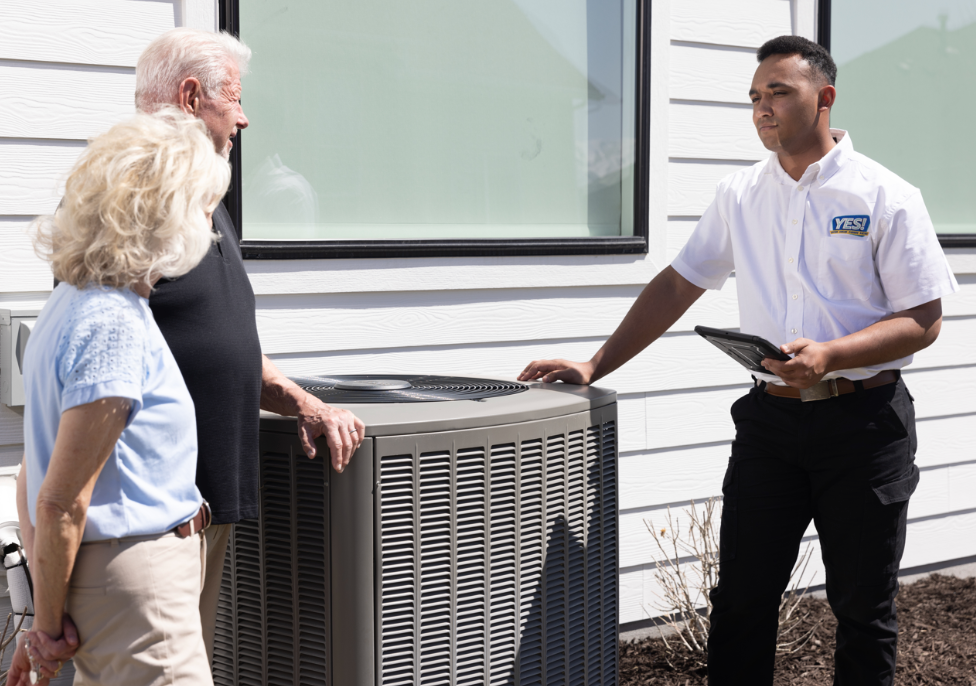 YES! technician in uniform with a tablet discussing an outdoor air conditioning unit with an elderly couple outside a white house on a sunny day.